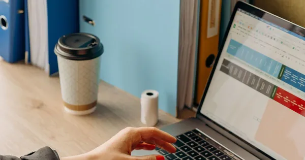 Manos sobre el teclado de un portátil que muestra una planilla, junto a un café