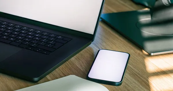 Teléfono con la pantalla en blanco junto a un computador portátil sobre un escritorio de madera