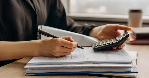 Manos revisando un documento timbrado con una calculadora, junto a un archivador de papeles sobre un escritorio